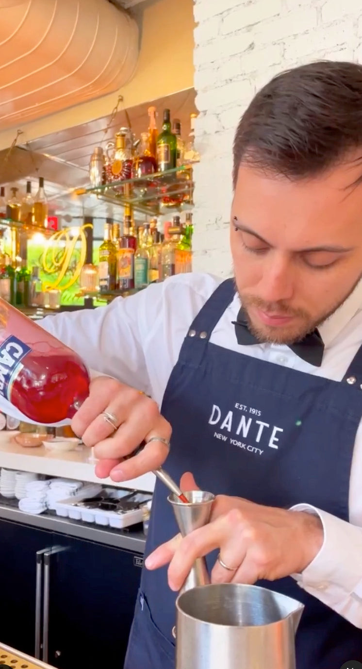 Bartender at Dante New York wearing a smart-casual uniform with denim apron and bowtie, emphasizing casual approachability.