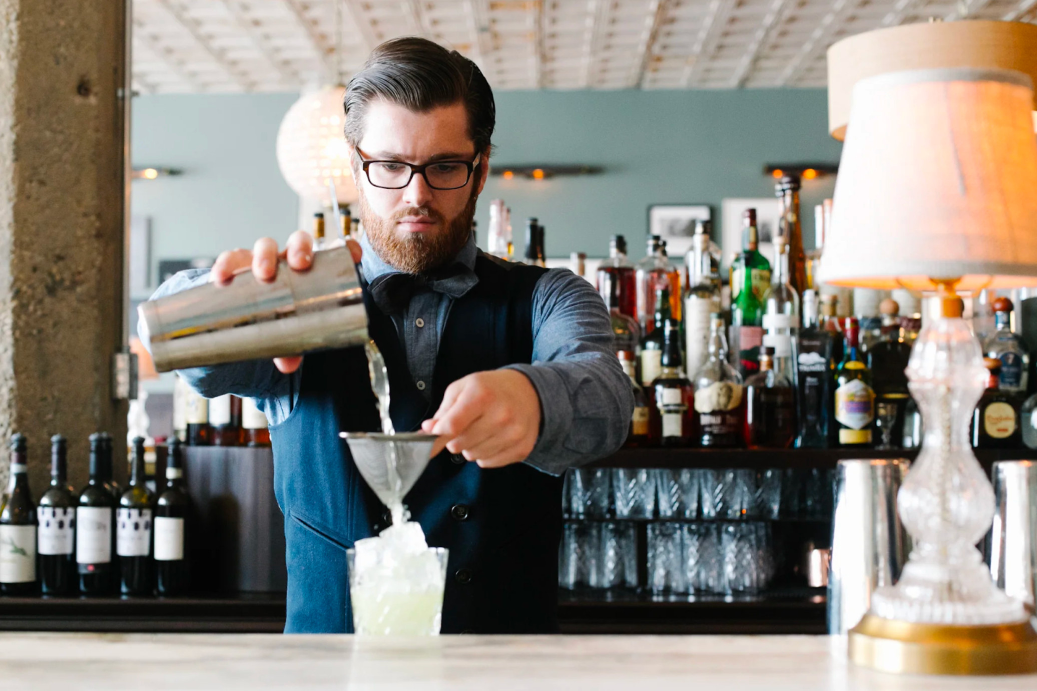 Bartender at Soho House Chicago in a chambray club collar shirt, twill vest, corduroy bow tie, and herringbone trousers, showcasing refined British-American style.